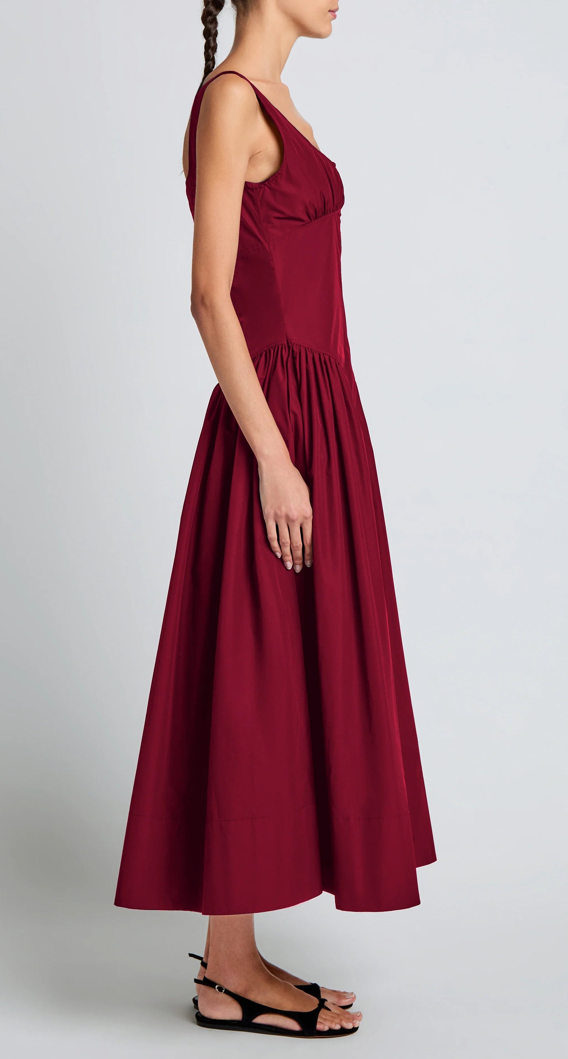 A woman in a sleeveless, ankle-length burgundy Kendall Dress by Proenza Schouler White Label, featuring a scoop neckline and crafted from soft Tech Cotton, stands in profile against a plain light background wearing black sandals.