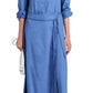 A person is wearing the Proenza Schouler White Label Lato Wrap Dress Linen Slub, a long blue button-up style with a shirtdress silhouette and belt, holding a white clutch. The sleeves are rolled up, and they complement it with black sandals for a chic finish.