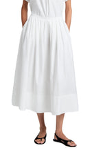 A person wearing the Proenza Schouler White Label Austin Skirt in white cotton sateen, knife-pleated and high-waisted, with hands in pockets, paired with black sandals.
