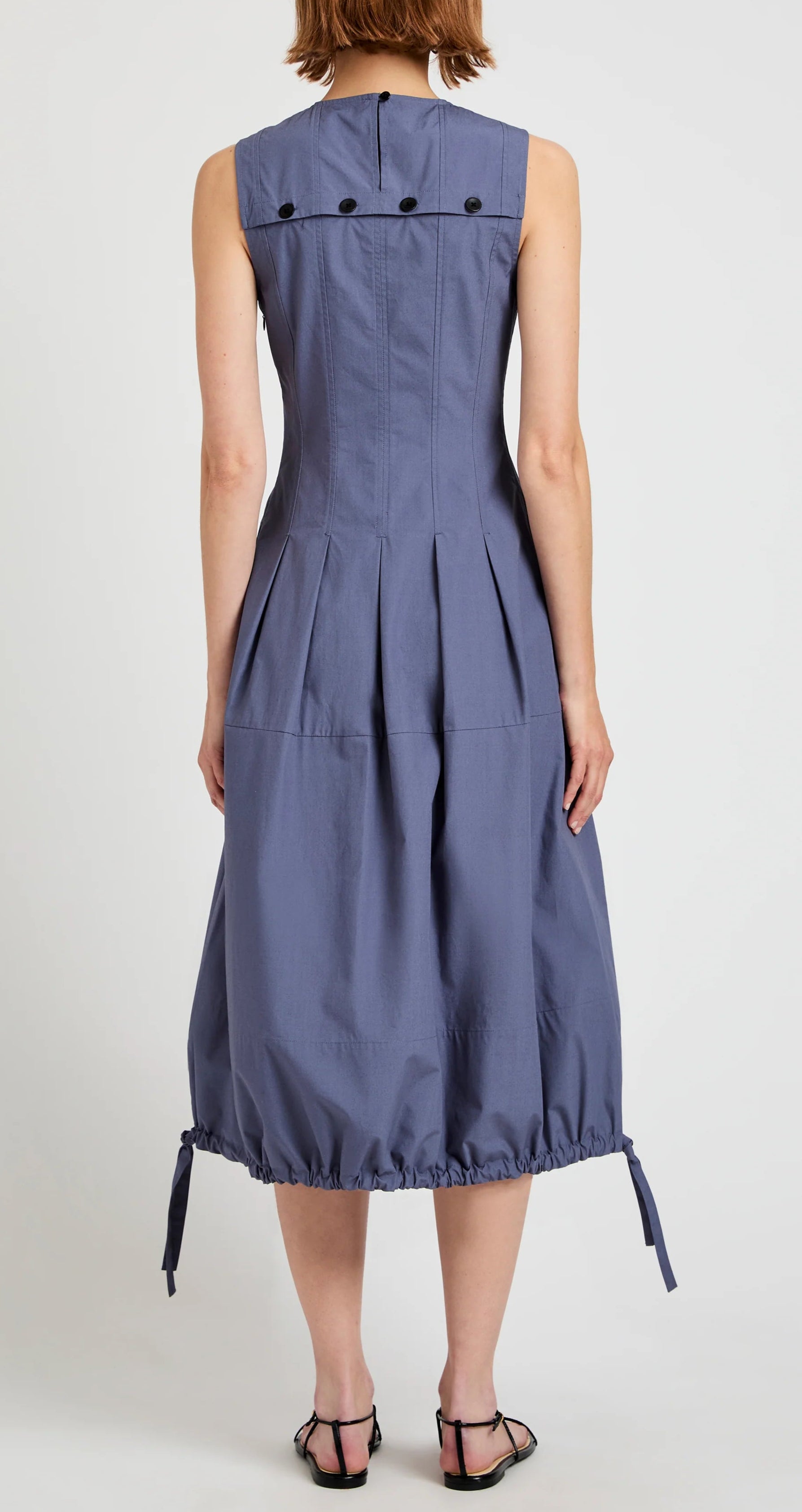 A woman stands with her back to the camera, wearing the Marley Brushed Poplin Dress by Proenza Schouler White Label—a sleeveless blue A-line midi with black buttons, pleats, and a gathered bubble hem with ties.