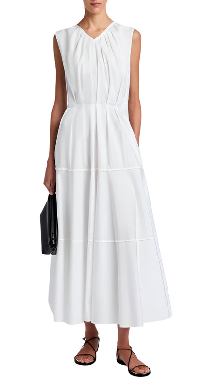 A woman wears the Kailey Dress Stretch Poplin by Proenza Schouler White Label—a sleeveless white V-neck pleated dress—paired with black strappy sandals and a black clutch.