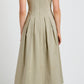 A person wearing the Proenza Schouler White Label Shelby Dress, a sleeveless beige cotton fit-and-flare midi with pleats, seen from the back; black flats and a visible black strap complete the look.