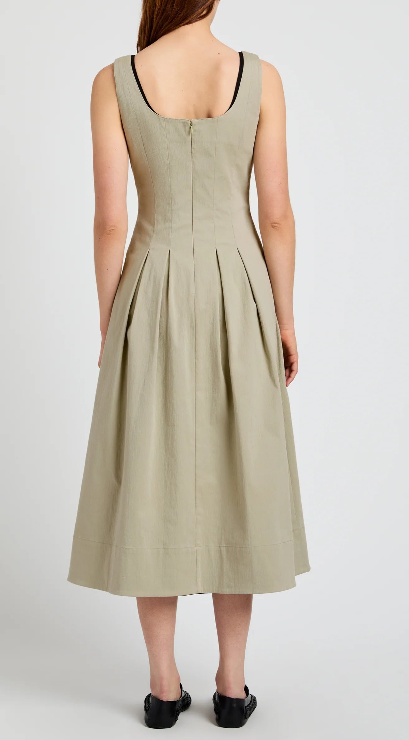 A person wearing the Proenza Schouler White Label Shelby Dress, a sleeveless beige cotton fit-and-flare midi with pleats, seen from the back; black flats and a visible black strap complete the look.
