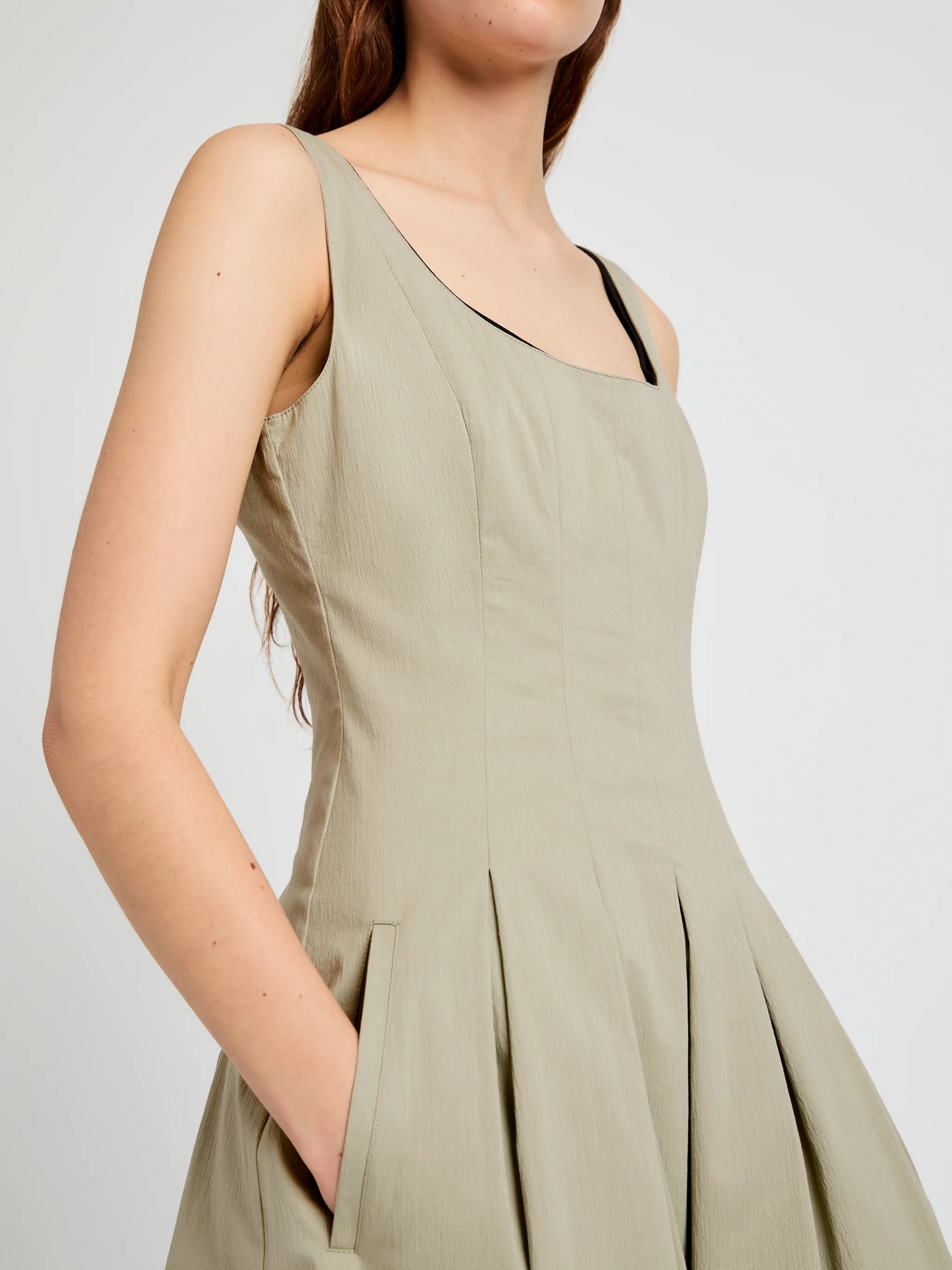 A person wears the Shelby Dress by Proenza Schouler White Label—a sleeveless, light beige fit-and-flare style with pleats, pockets, and a square neckline—standing against a plain light background.