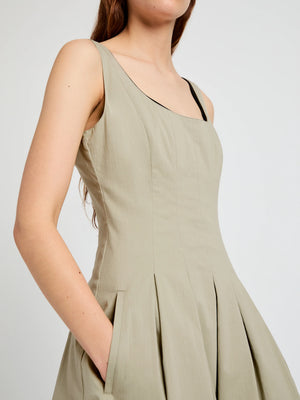 A person wears the Shelby Dress by Proenza Schouler White Label—a sleeveless, light beige fit-and-flare style with pleats, pockets, and a square neckline—standing against a plain light background.