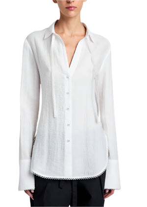 A woman wears the Proenza Schouler White Label Cami Shirt Crinkle Viscose in white, a relaxed-fit top with a collar, long sleeves, and subtle trim at the hem, paired with black pants.