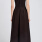A woman faces away in the Proenza Schouler White Label Bronwyn Merino Wool Twill Dress, a sleeveless dark brown A-line style with pleat tuck details, paired with black pointed boots.