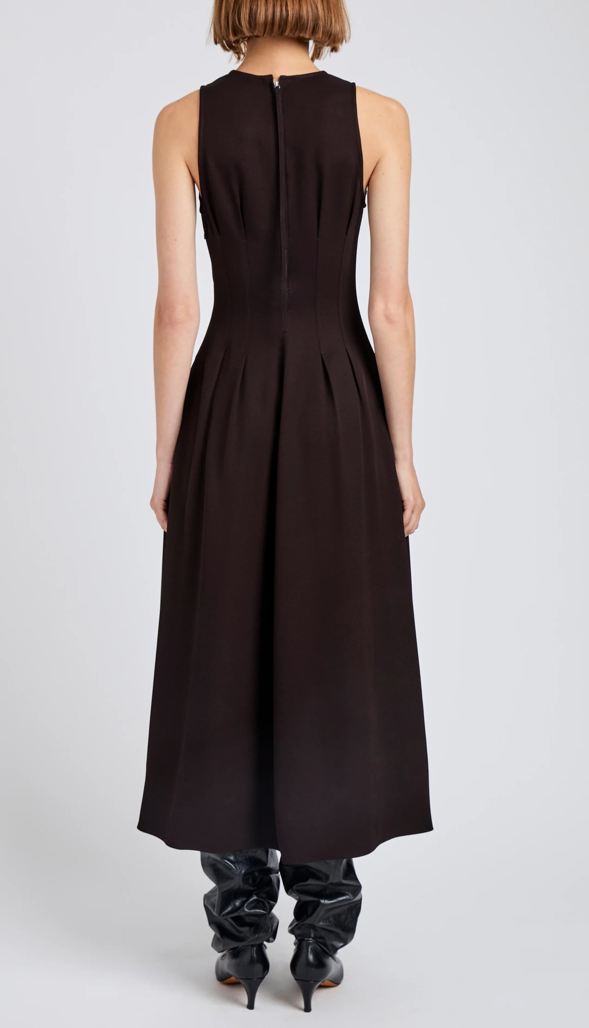 A woman faces away in the Proenza Schouler White Label Bronwyn Merino Wool Twill Dress, a sleeveless dark brown A-line style with pleat tuck details, paired with black pointed boots.