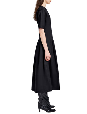 A person in the Proenza Schouler White Label Ainsley Dress in black tech cotton, featuring an A-line silhouette, short sleeves, and a flared midi skirt, styled with black heeled boots and shown in side profile against a white background.
