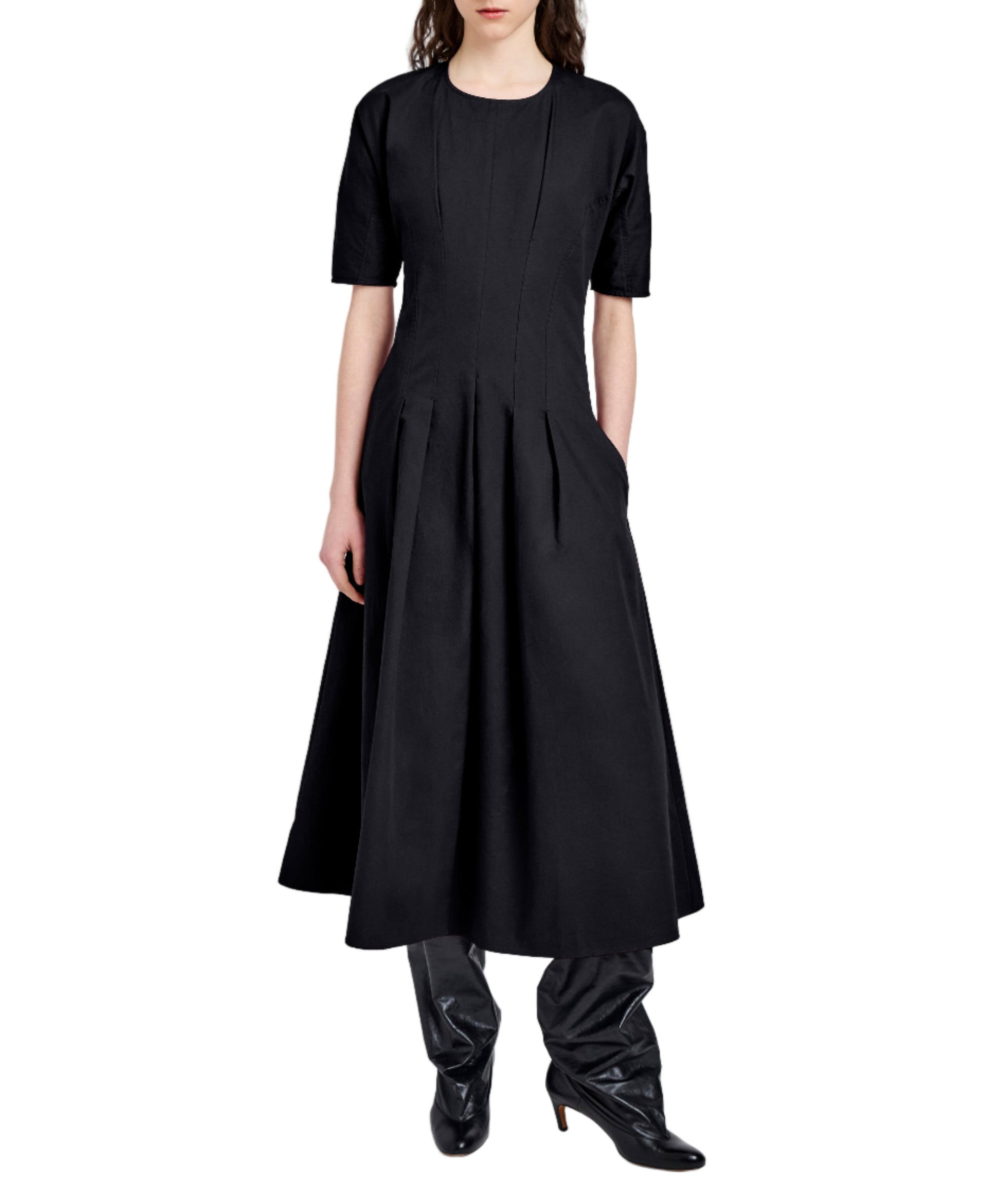 Someone wears the Ainsley Dress by Proenza Schouler White Label—a black, ankle-length, short-sleeve dress with pleats and an A-line silhouette—paired with black heeled boots.