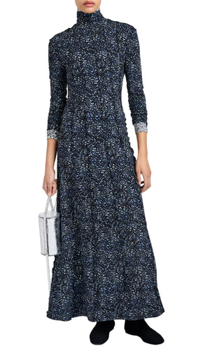 A person wears the Proenza Schouler White Label Mabel Cheetah Jersey Dress, a long-sleeve turtleneck maxi in dark blue pattern, styled with black shoes and a small metallic handbag.