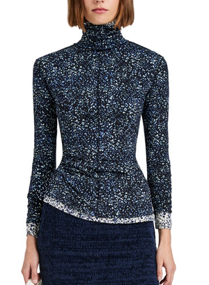 A woman wears the Proenza Schouler White Label Audrey Cheetah Jersey Top, a long-sleeve, turtleneck style with a subtle blue and white pattern, paired with a textured dark skirt. The image is cropped at the shoulders and thighs.