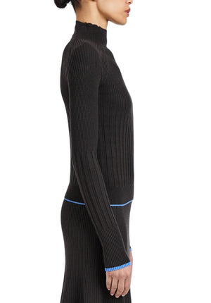 A woman in profile wears the Proenza Schouler White Label Daniella Sweater, a fitted ribbed black skirt set with blue trim at the waist and cuffs, standing against a white background.