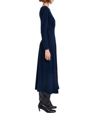 A model wears the Proenza Schouler White Label Sai Wool Jersey Dress—navy blue, long-sleeve, A-line with a V-cut back—paired with black heeled boots, shown in profile against a white background.