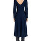 A woman wearing the Proenza Schouler White Label Sai Wool Jersey Dress—a navy blue, long-sleeve A-line midi with a V-cut back—paired with black heeled boots, is shown from behind against a white background.