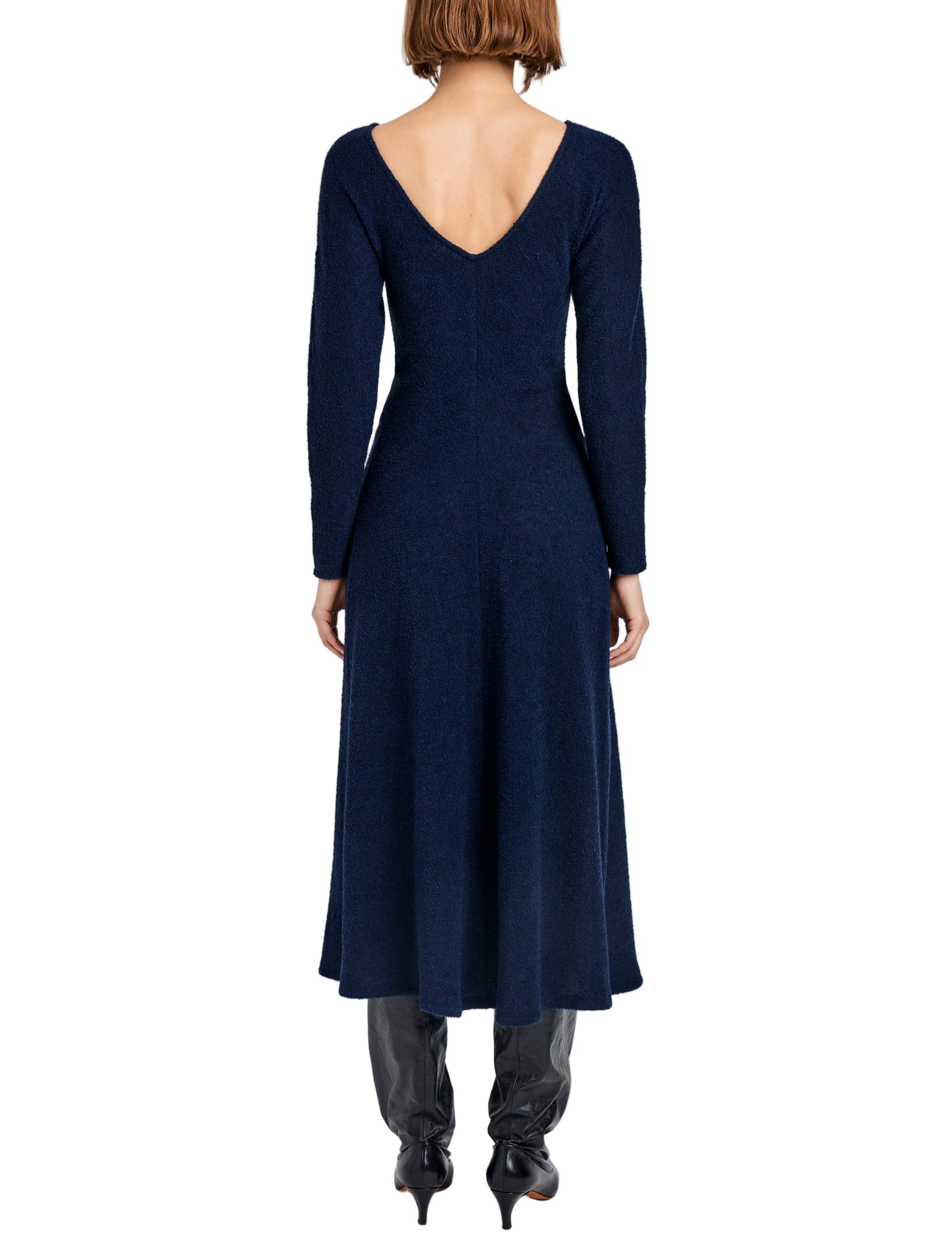 A woman wearing the Proenza Schouler White Label Sai Wool Jersey Dress—a navy blue, long-sleeve A-line midi with a V-cut back—paired with black heeled boots, is shown from behind against a white background.