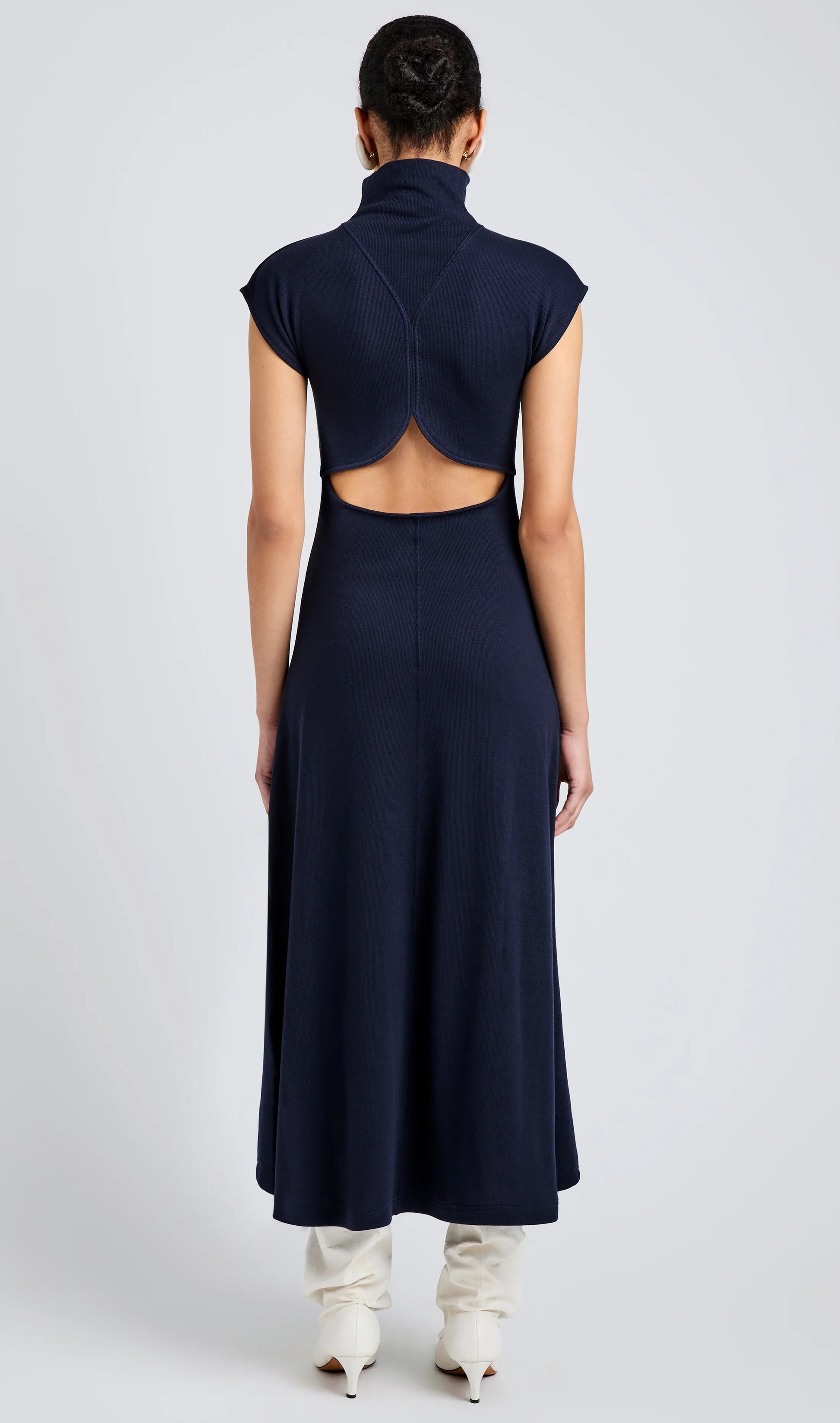 A person faces away, wearing the Proenza Schouler White Label Brea Brushed Rib Dress—a navy sleeveless A-line design with a high collar and circular back cutout—paired with white boots.
