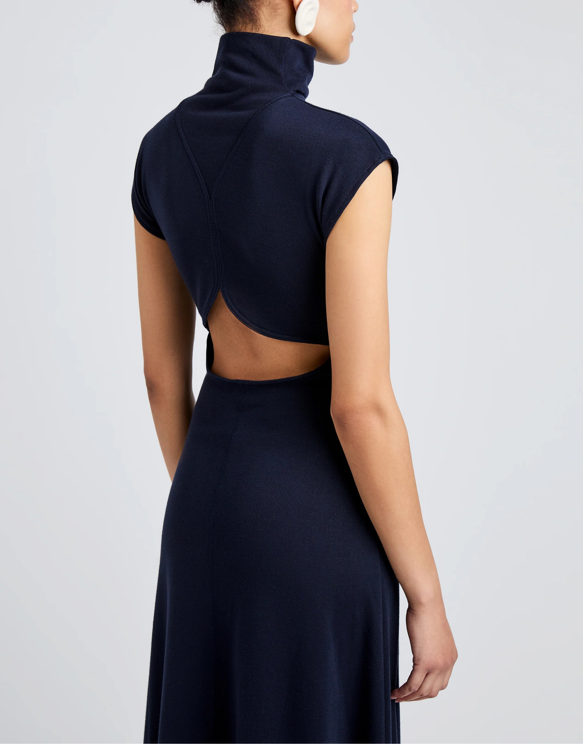 A person wears the Proenza Schouler White Label Brea Brushed Rib Dress in navy, featuring a high collar, short sleeves, open back, and A-line silhouette. The figure is seen from behind against a plain light background.