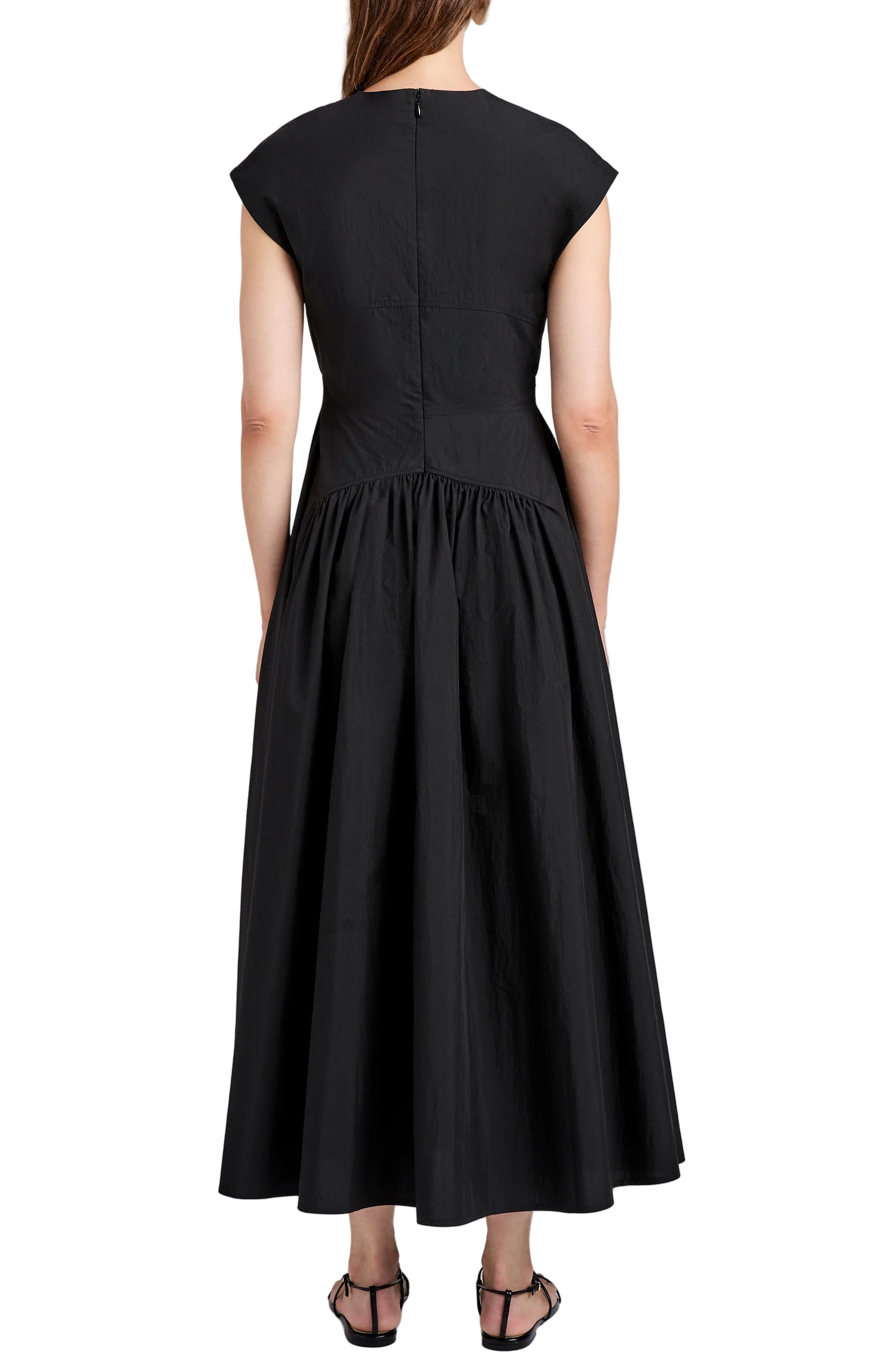 A woman faces away in the Proenza Schouler White Label Nyssa Dress—an ankle-length, black tech cotton piece with short sleeves, A-line silhouette, and gathered skirt—paired with black sandals against a plain background.