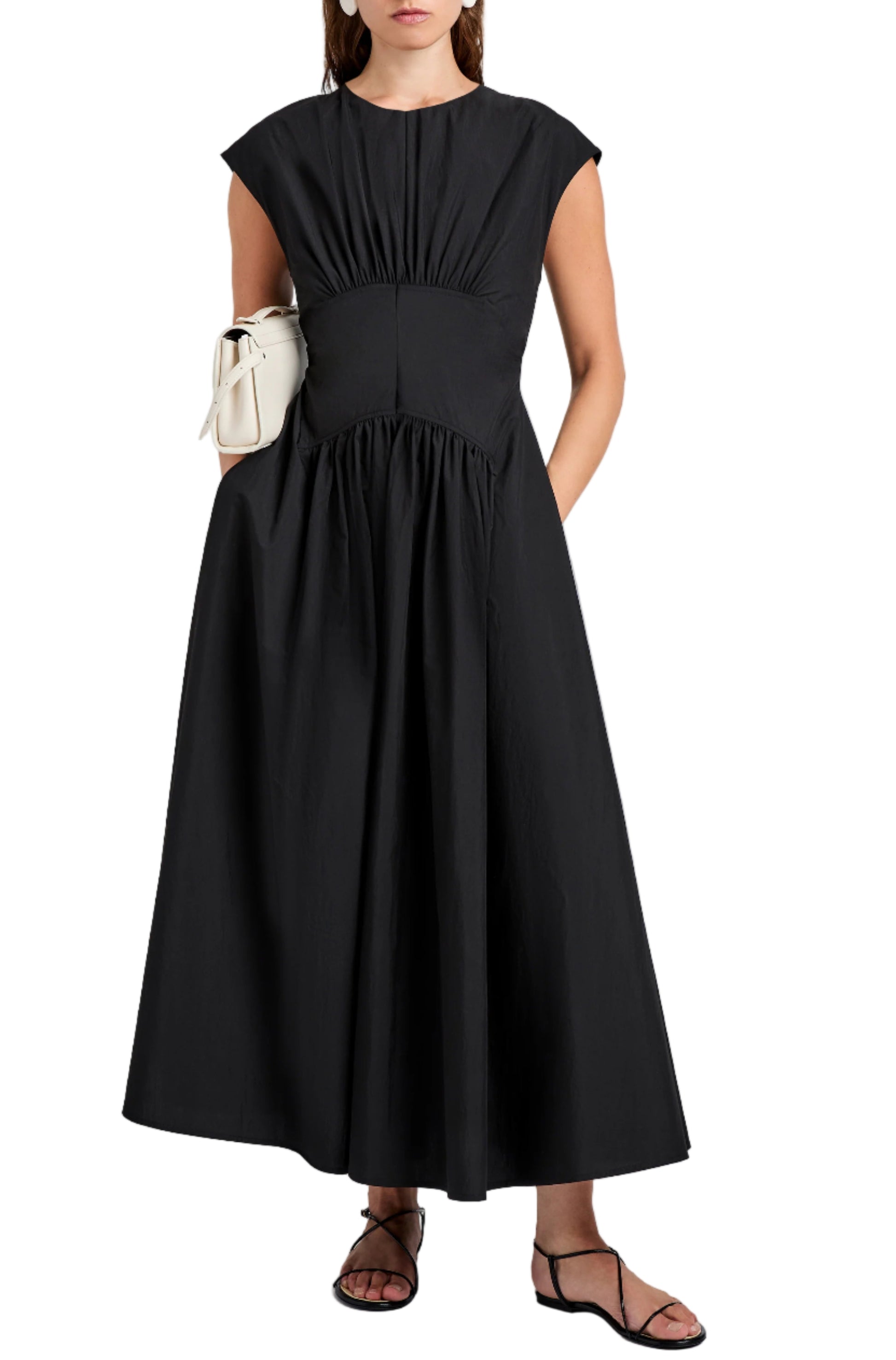 A woman wears the Nyssa Dress by Proenza Schouler White Label—an ankle-length, sleeveless black A-line dress with shirring—paired with a white clutch and black sandals.