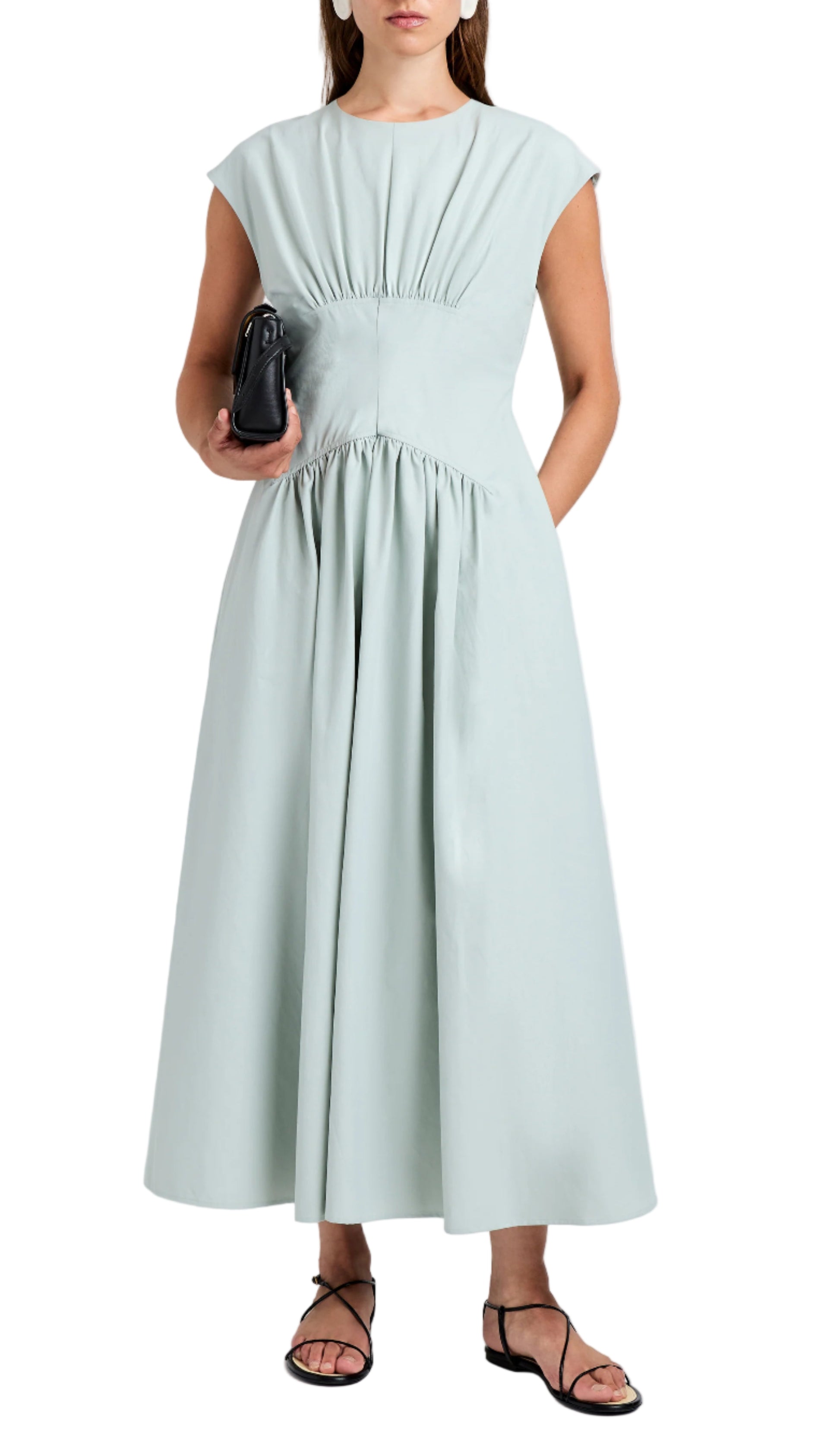 A woman wears the Proenza Schouler White Label Nyssa Dress, a light blue sleeveless midi with shirring and an A-line silhouette, styled with black sandals and a black clutch.