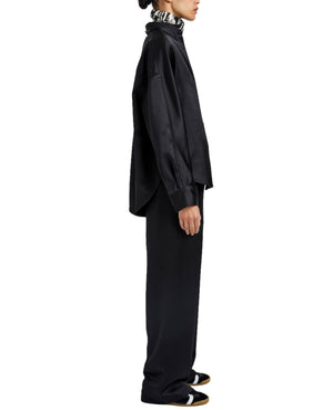 A person stands in profile wearing the Proenza Schouler White Label Caroline Crinkled Crepe Shirt, a loose black top, black wide-leg pants for a relaxed silhouette, and black shoes with white and brown details.