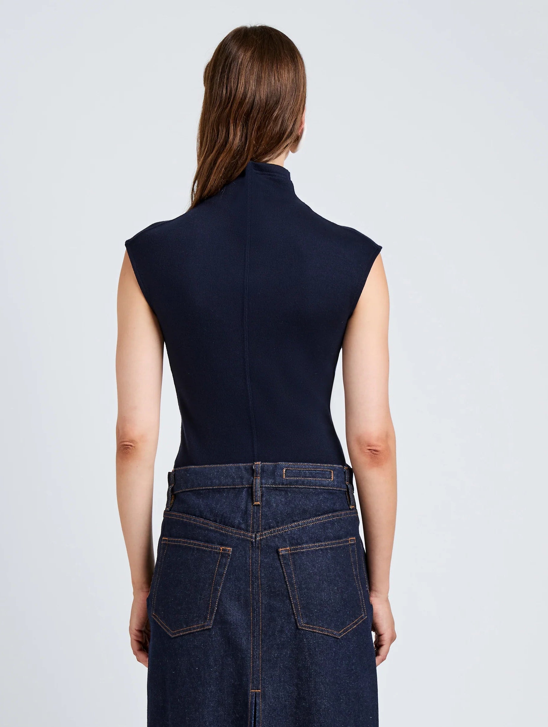 A person with long brown hair faces away from the camera, wearing the Proenza Schouler White Label Zane Brushed Rib Top and a high-waisted dark denim skirt.