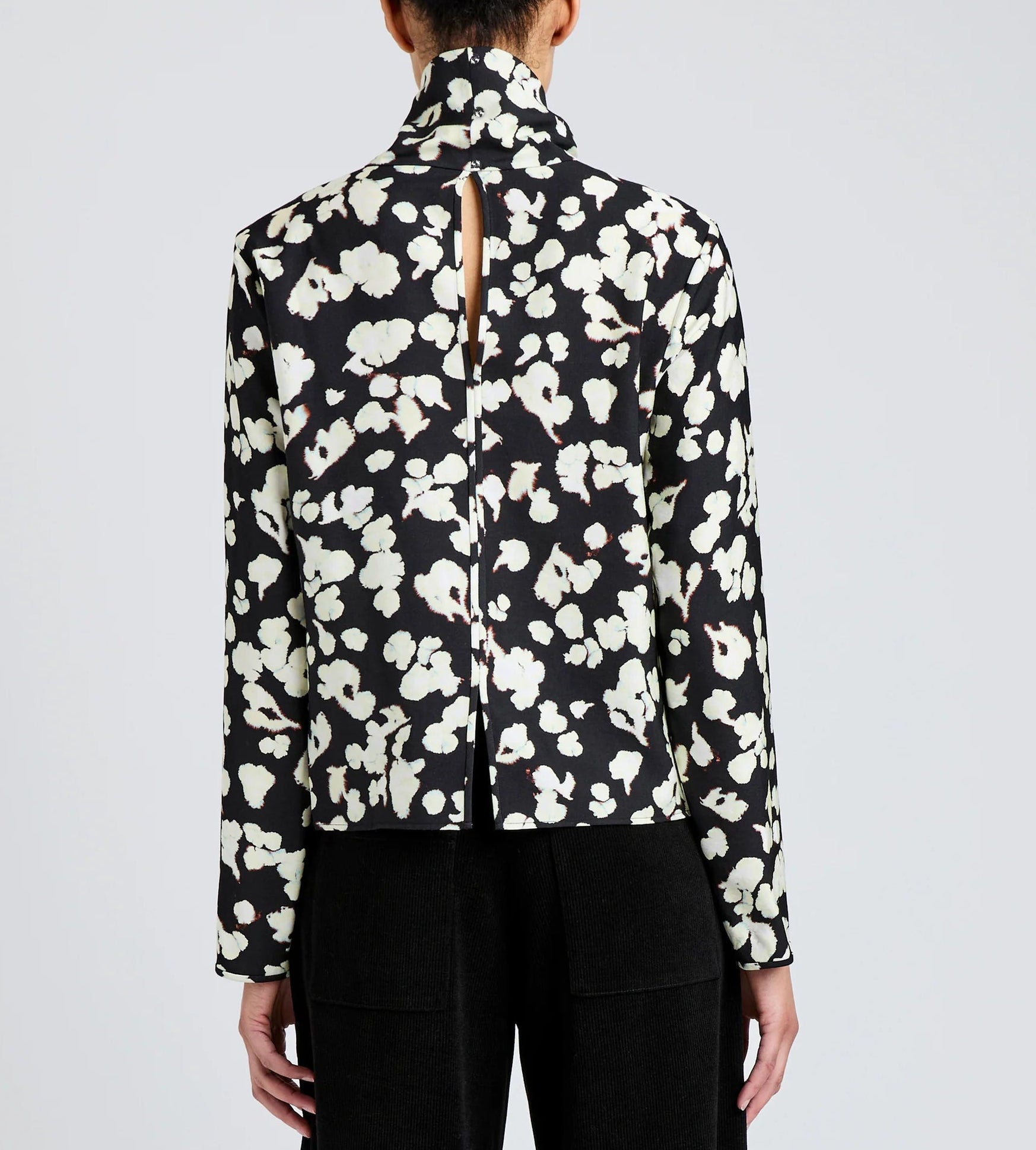 Person wearing the Ansel Top by Proenza Schouler White Label, a black long-sleeve with a white abstract floral print and vertical back slit, paired with black pants.