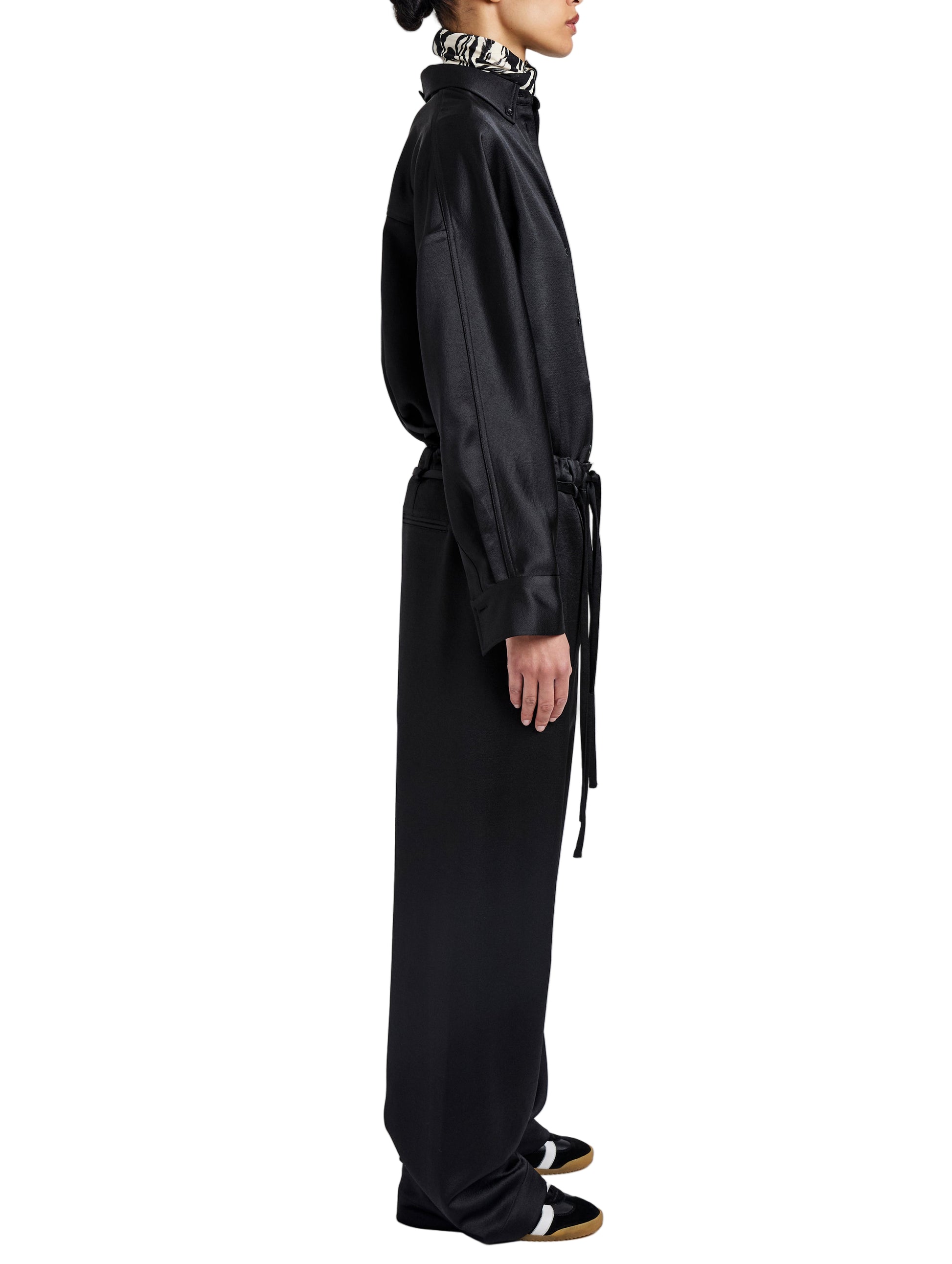 A woman in profile wears the Proenza Schouler White Label Magnus Crinkled Crepe Pant—long, black, high-waisted, wide-leg—with a belted waist, black and white shoes, and a patterned scarf or collar.