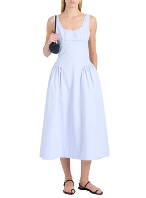 A woman wears the Proenza Schouler White Label Kendall Dress, a sleeveless light blue tech cotton midi with a scoop neckline, paired with black sandals and a small black shoulder bag.