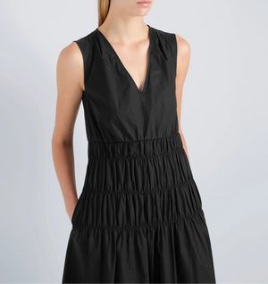 A woman wears the Proenza Schouler White Label Loren Dress—a sleeveless black V-neck poplin dress with an A-line silhouette and gathered, tiered skirt—standing against a plain light background.
