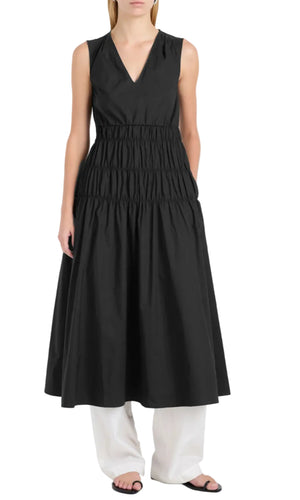 A person wears the sleeveless black Loren Dress by Proenza Schouler White Label, featuring a V-neck and gathered details in airy poplin, over loose white pants and black sandals, standing against a white background.