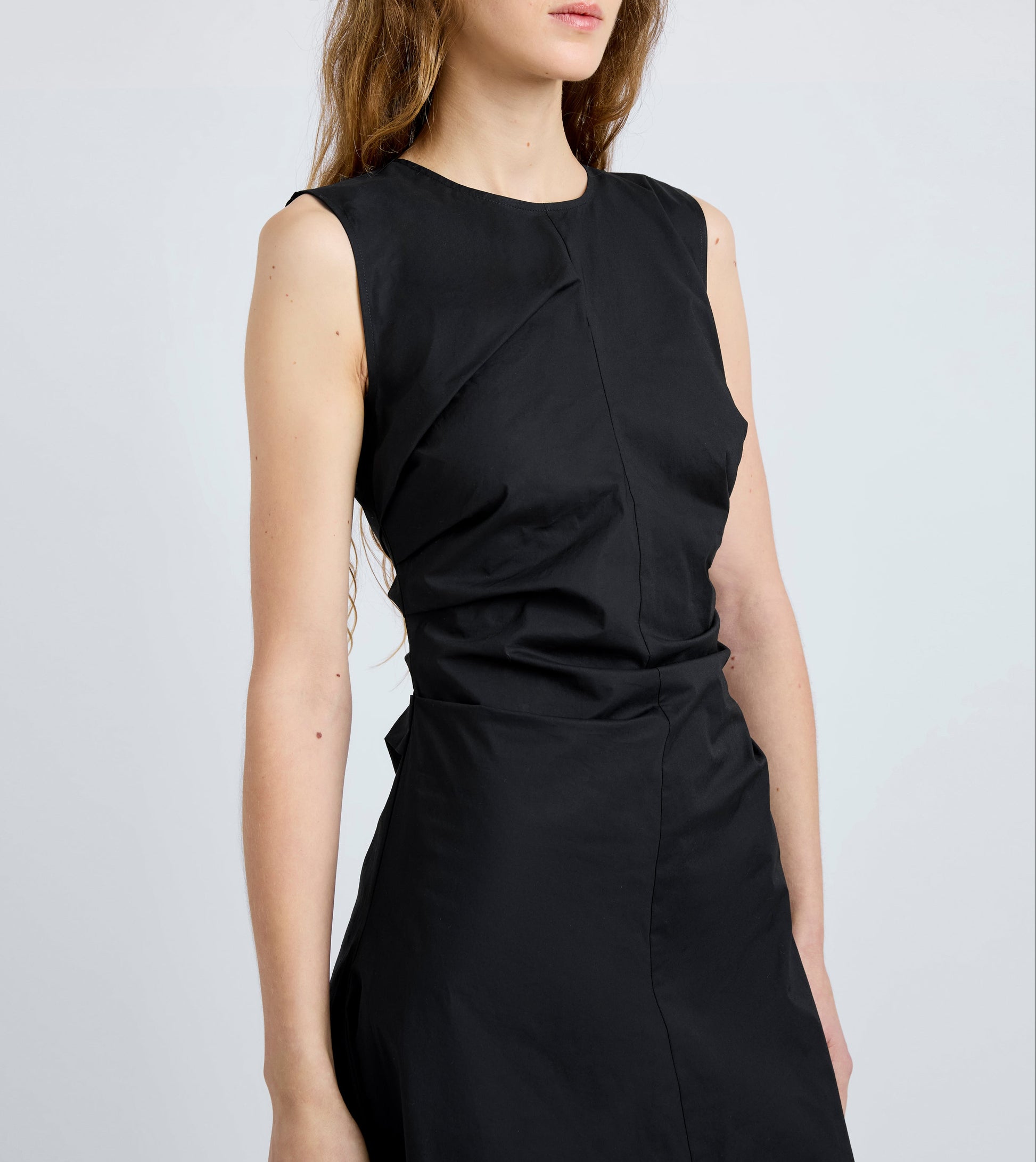 A woman in the sleeveless black Anoushka Dress by Proenza Schouler White Label stands against a plain light background, her arms relaxed at her sides.