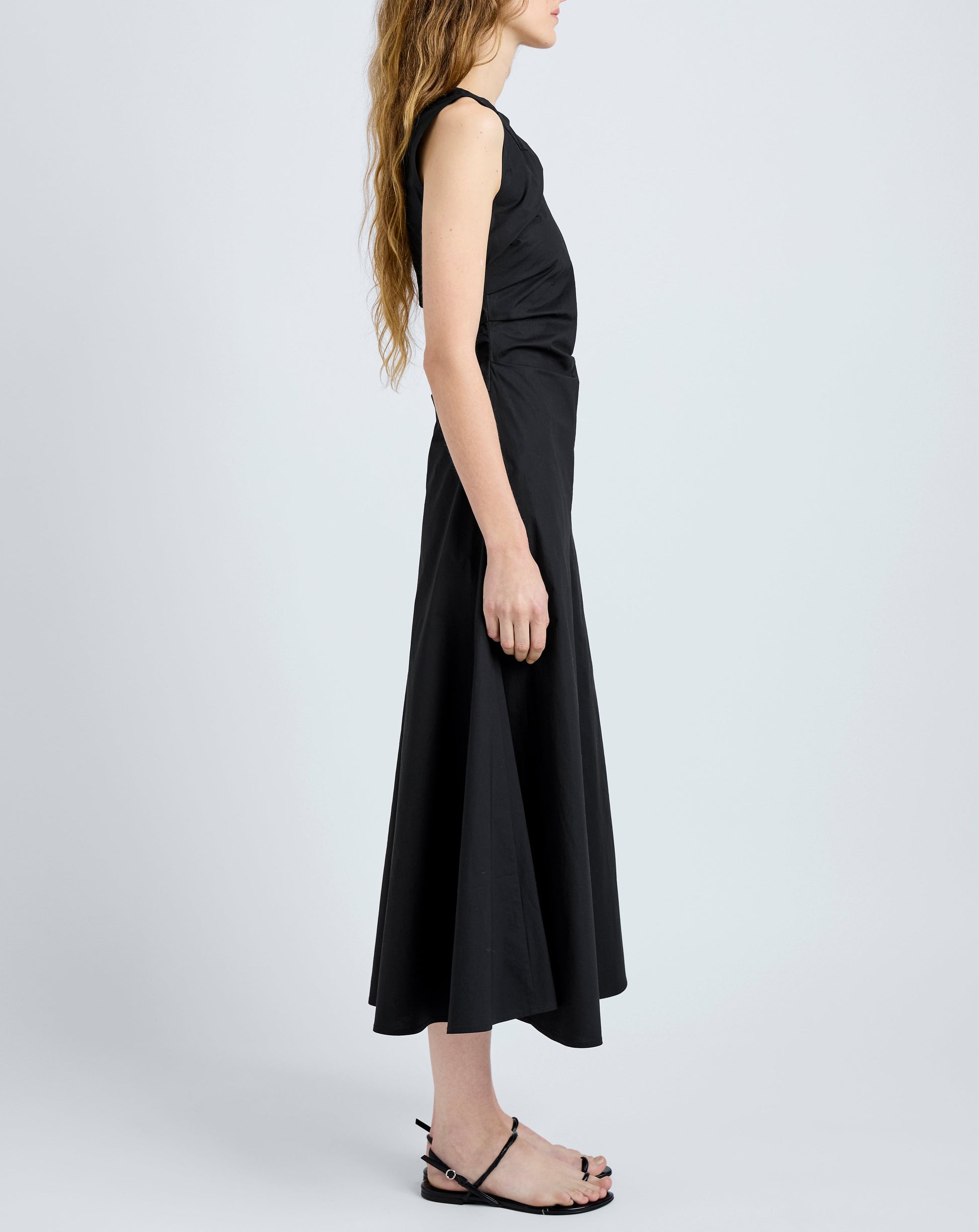 A woman stands in profile wearing the Proenza Schouler White Label Anoushka Dress, a sleeveless black midi poplin dress, paired with black flat sandals against a plain light background.
