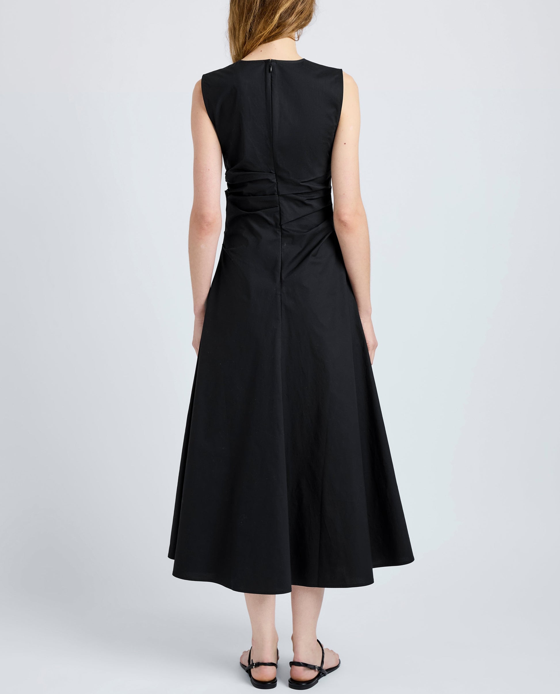 A woman, facing away, wears the Anoushka Dress by Proenza Schouler White Label—a sleeveless black poplin dress featuring a fitted bodice, flared skirt, back zipper, and subtle ruching—paired with black sandals.