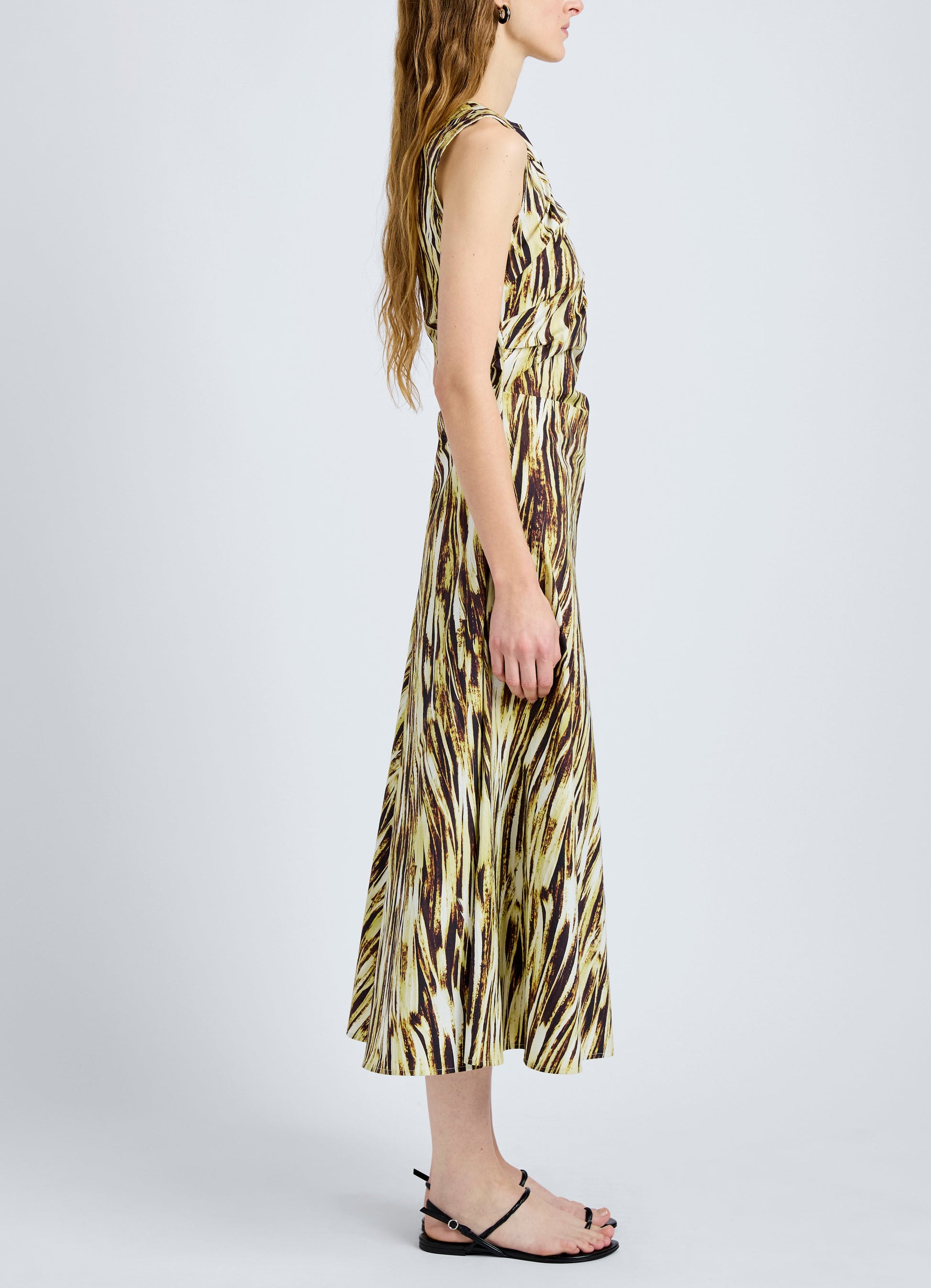 A woman stands in profile against a plain background, wearing the Proenza Schouler White Label Anoushka Dress, a sleeveless A-line style with a multicolored abstract print, paired with black sandals.