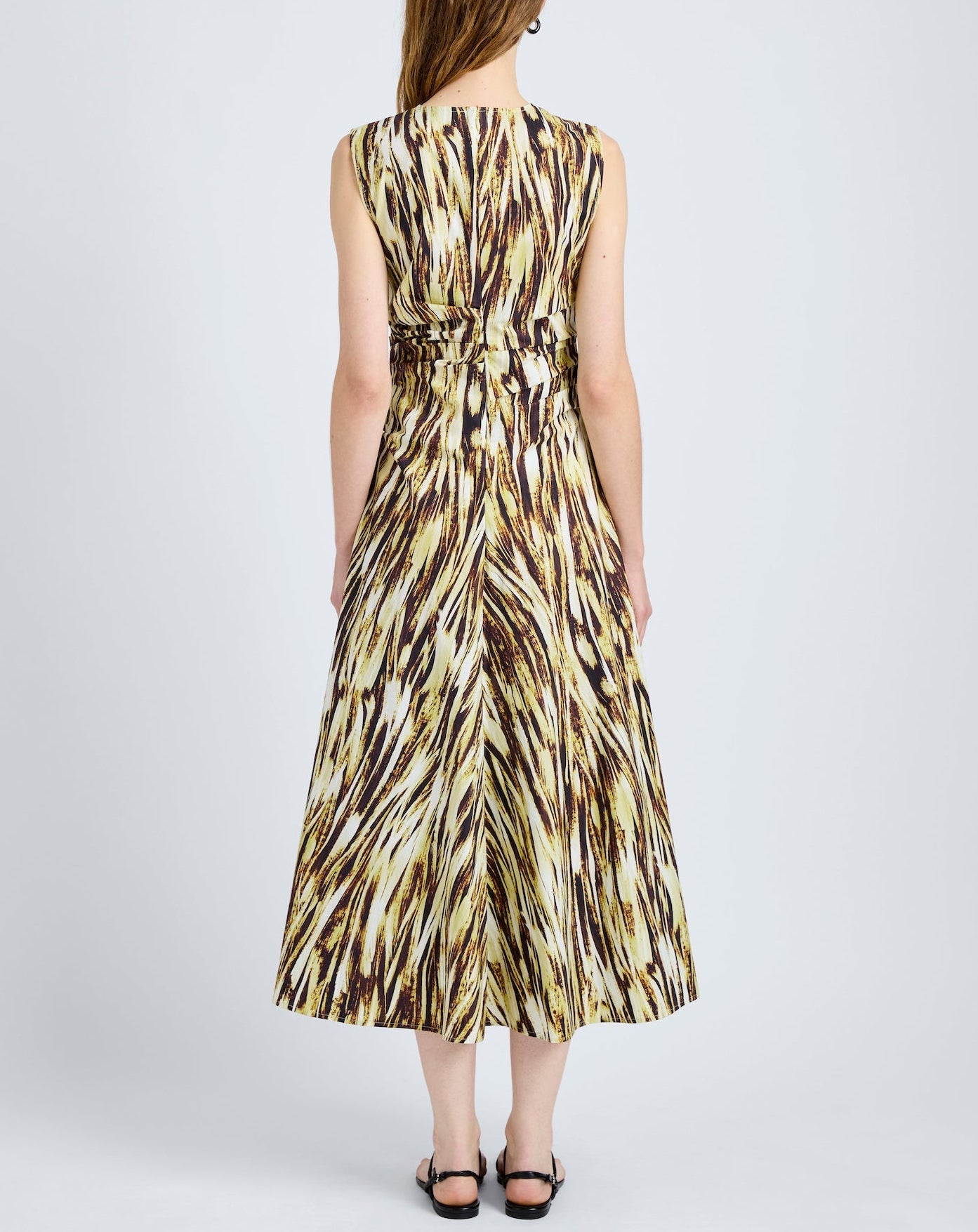 A woman stands facing away, wearing the Proenza Schouler White Label Anoushka Dress—a sleeveless, ankle-length poplin dress with a yellow, brown, and black abstract brushstroke print—paired with black sandals.