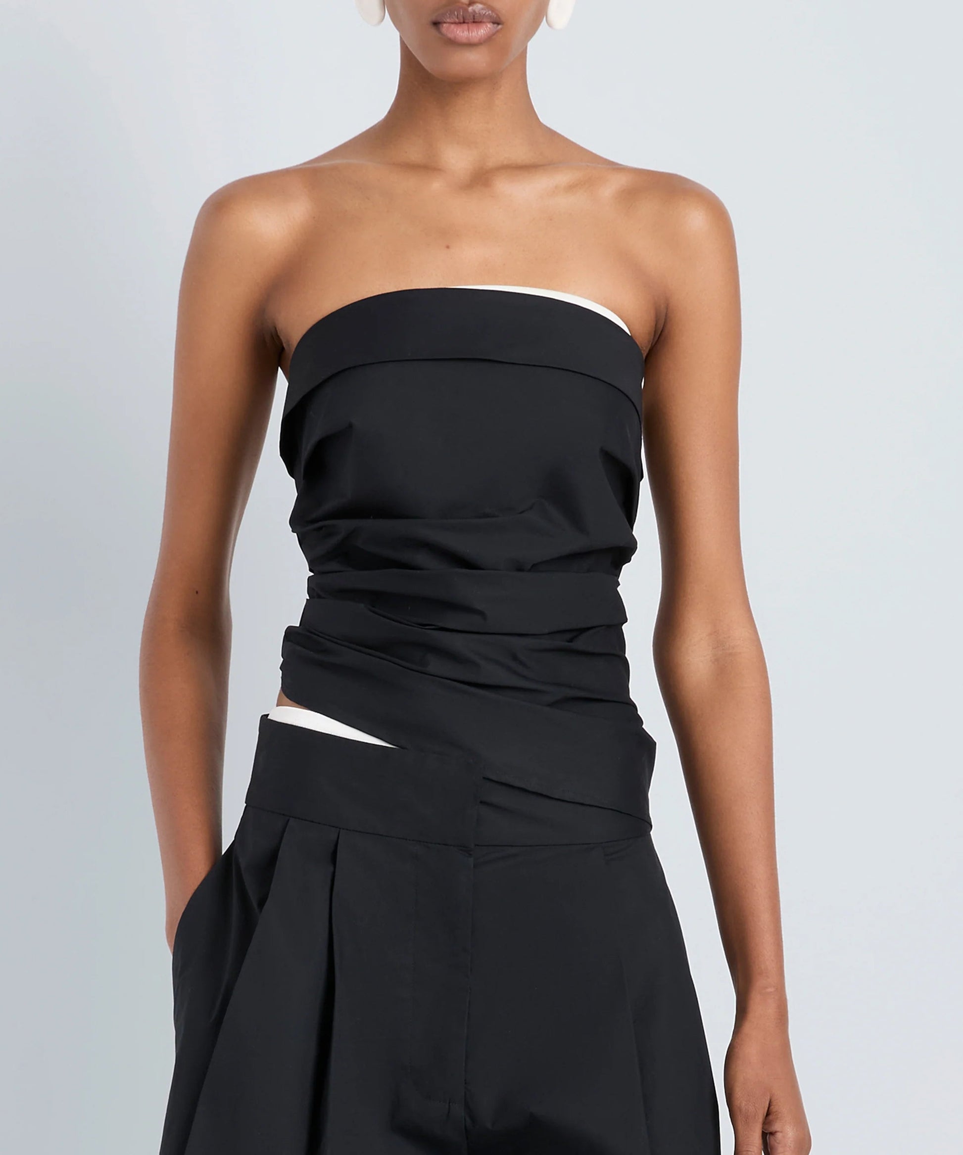 A person wearing the Maisy Strapless Top by Proenza Schouler White Label with black high-waisted pants, standing against a plain light gray background.