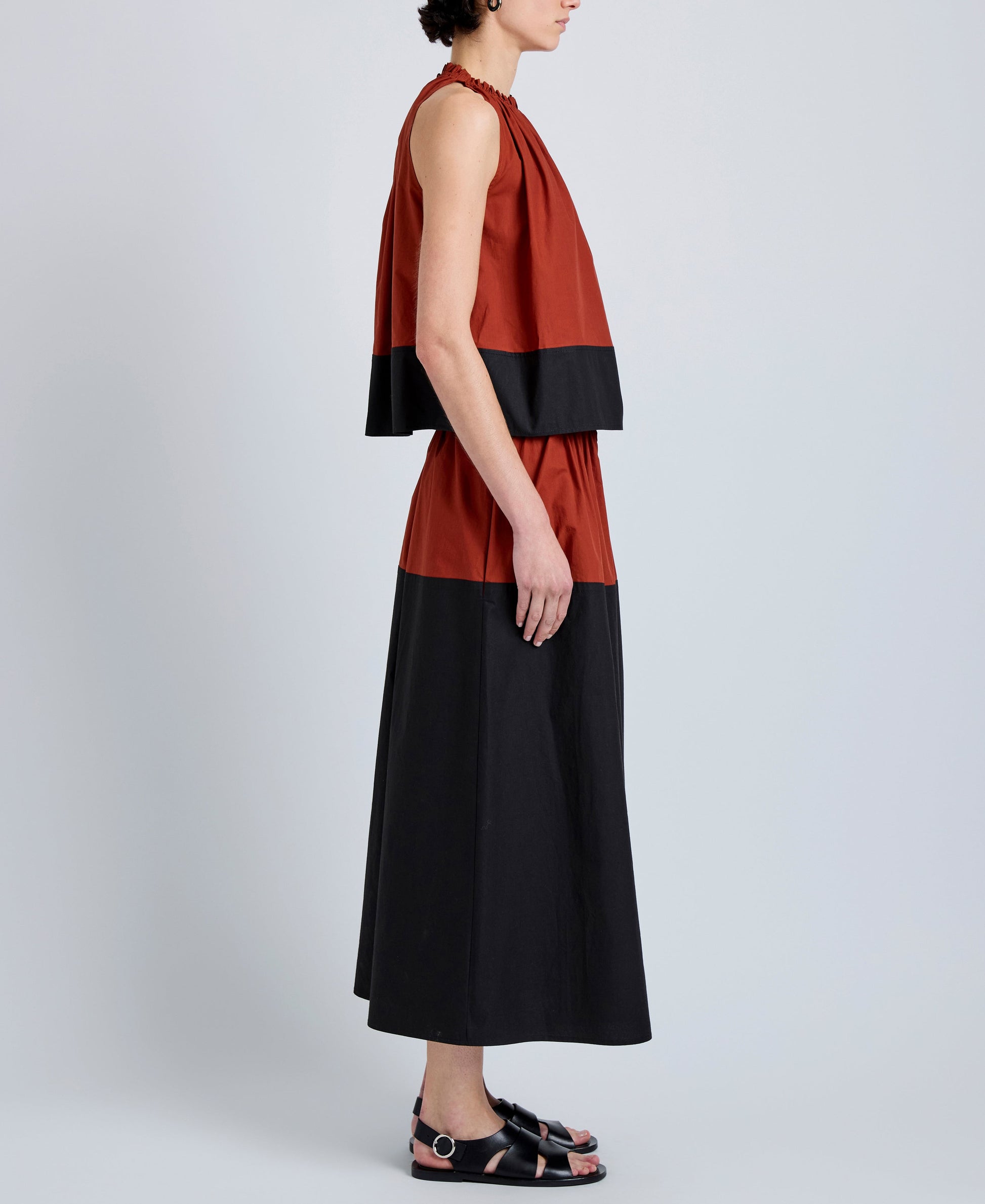 A person stands sideways against a plain background wearing the Esme Top by Proenza Schouler White Label in a cropped, sleeveless, rust and black color-blocked style, paired with black sandals.