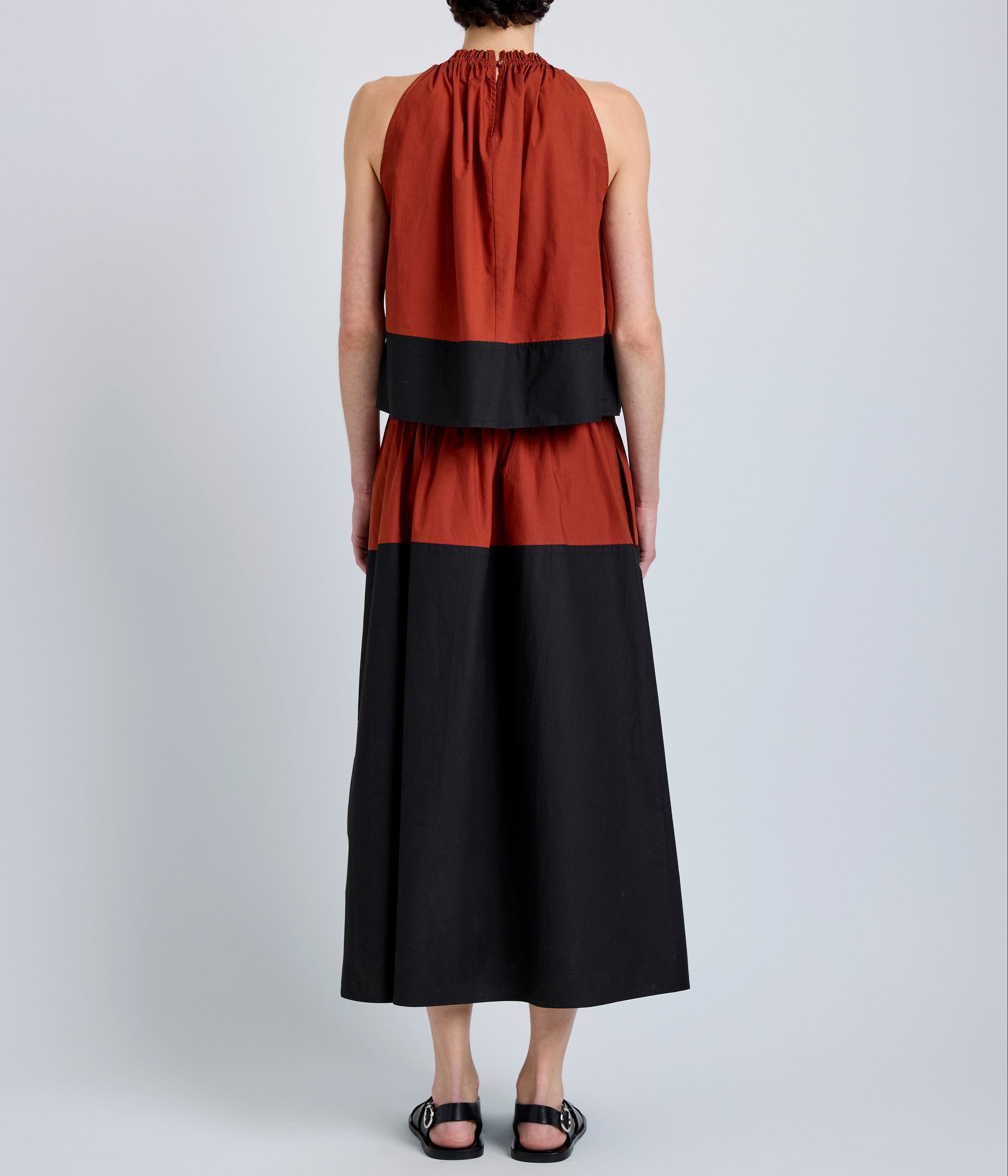 Person stands facing away, wearing the Esme Top by Proenza Schouler White Label in rust and black with a cropped silhouette, paired with black sandals, against a plain light gray background.
