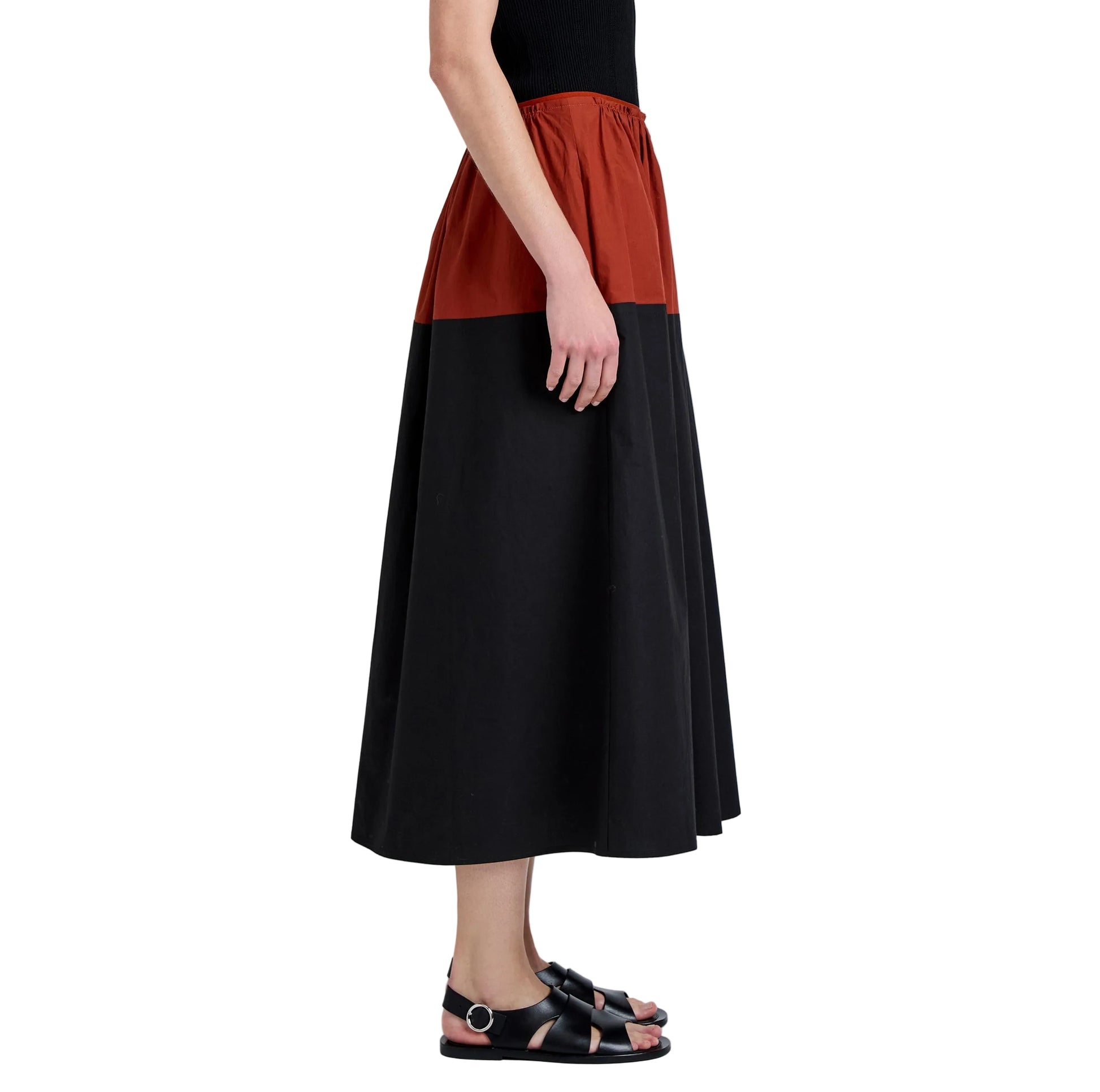 A person stands in profile wearing a sleeveless black top, the Nyla Skirt by Proenza Schouler White Label in color-blocked black and rust poplin, and black sandals against a white background.