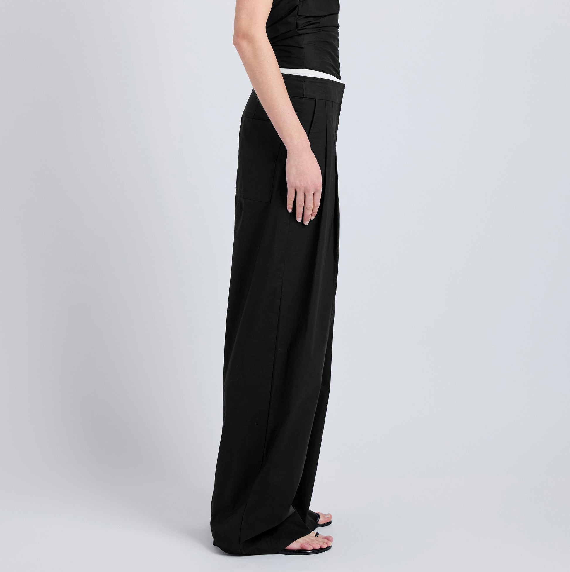 A person stands in profile against a plain light background, wearing the Coralie Pant by Proenza Schouler White Label and a black sleeveless top.