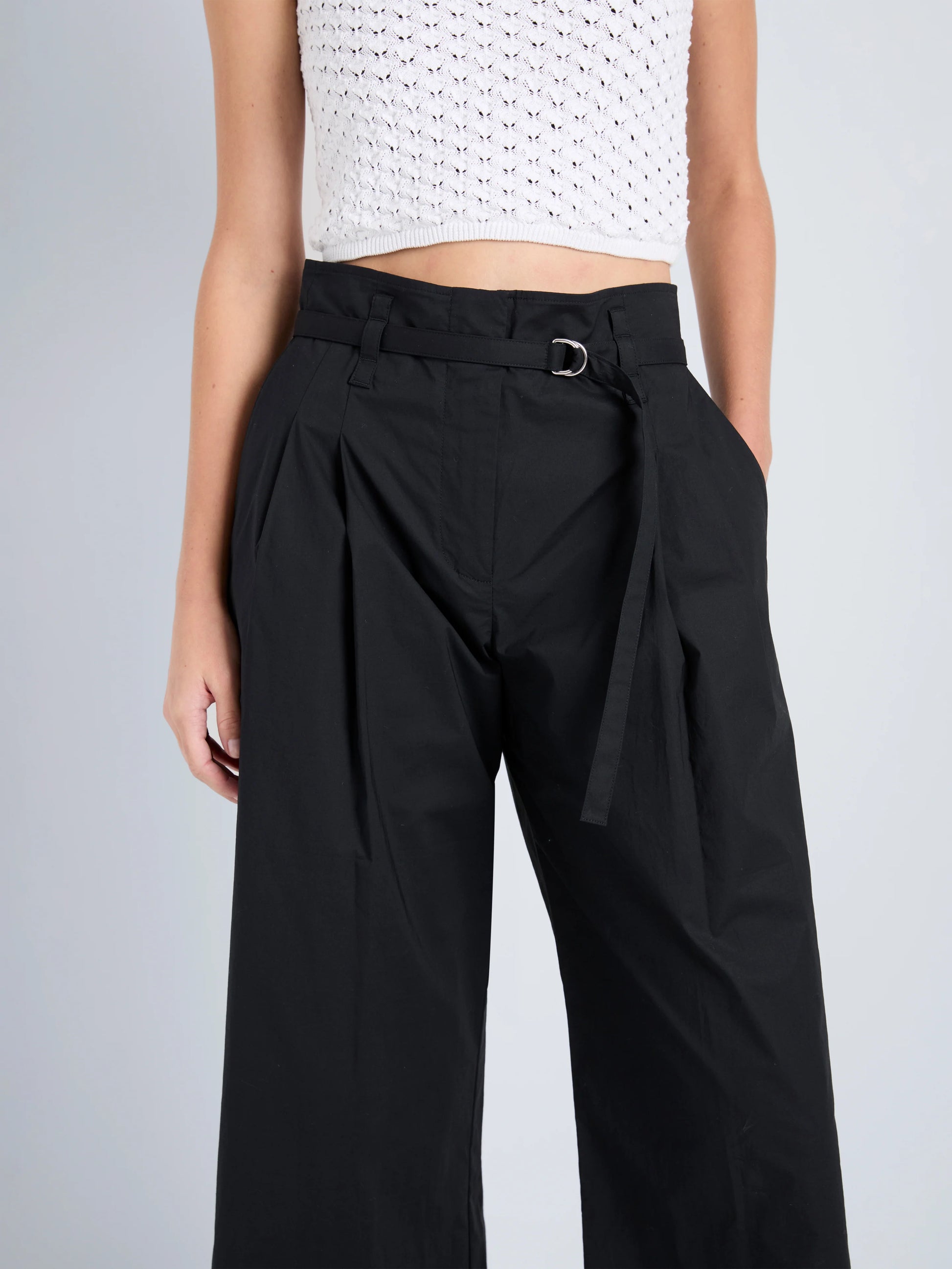 Model wears Proenza Schouler White Label Cropped Raver Pant in high-rise black with belt detail and a white crochet sleeveless crop top, posed against a plain background.