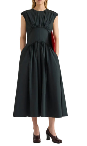 A woman wears the Proenza Schouler White Label Nyssa Stretch Cotton Dress—a sleeveless, gathered black dress featuring convenient pockets—styled with a red clutch and black heeled shoes.