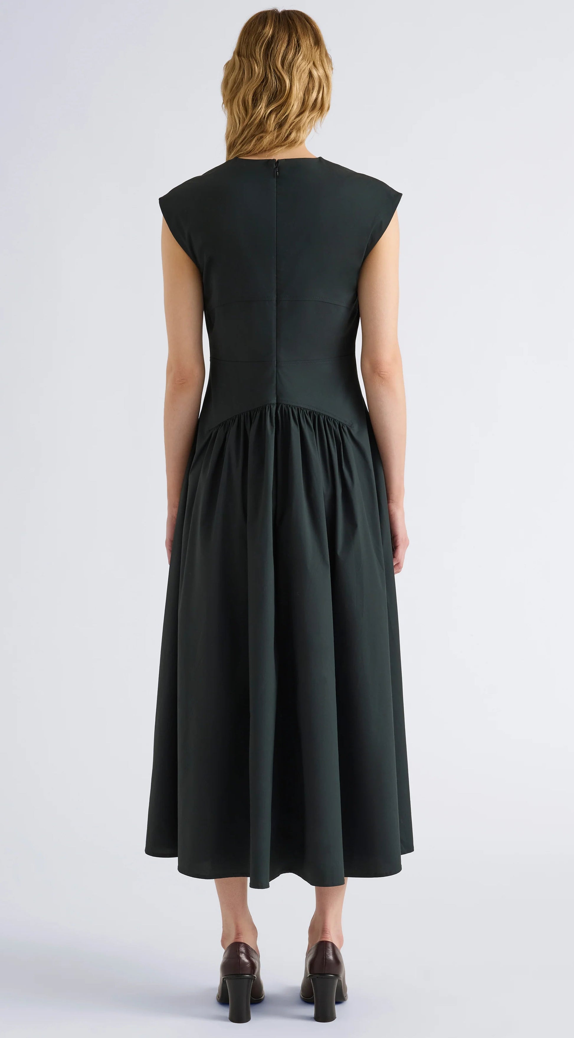 A woman stands facing away, wearing the Proenza Schouler White Label Nyssa Stretch Cotton Dress—a dark green, short-sleeved, midi-length dress for women featuring a fitted bodice and gathered skirt crafted from soft stretch cotton—stylishly paired with brown block-heeled shoes.