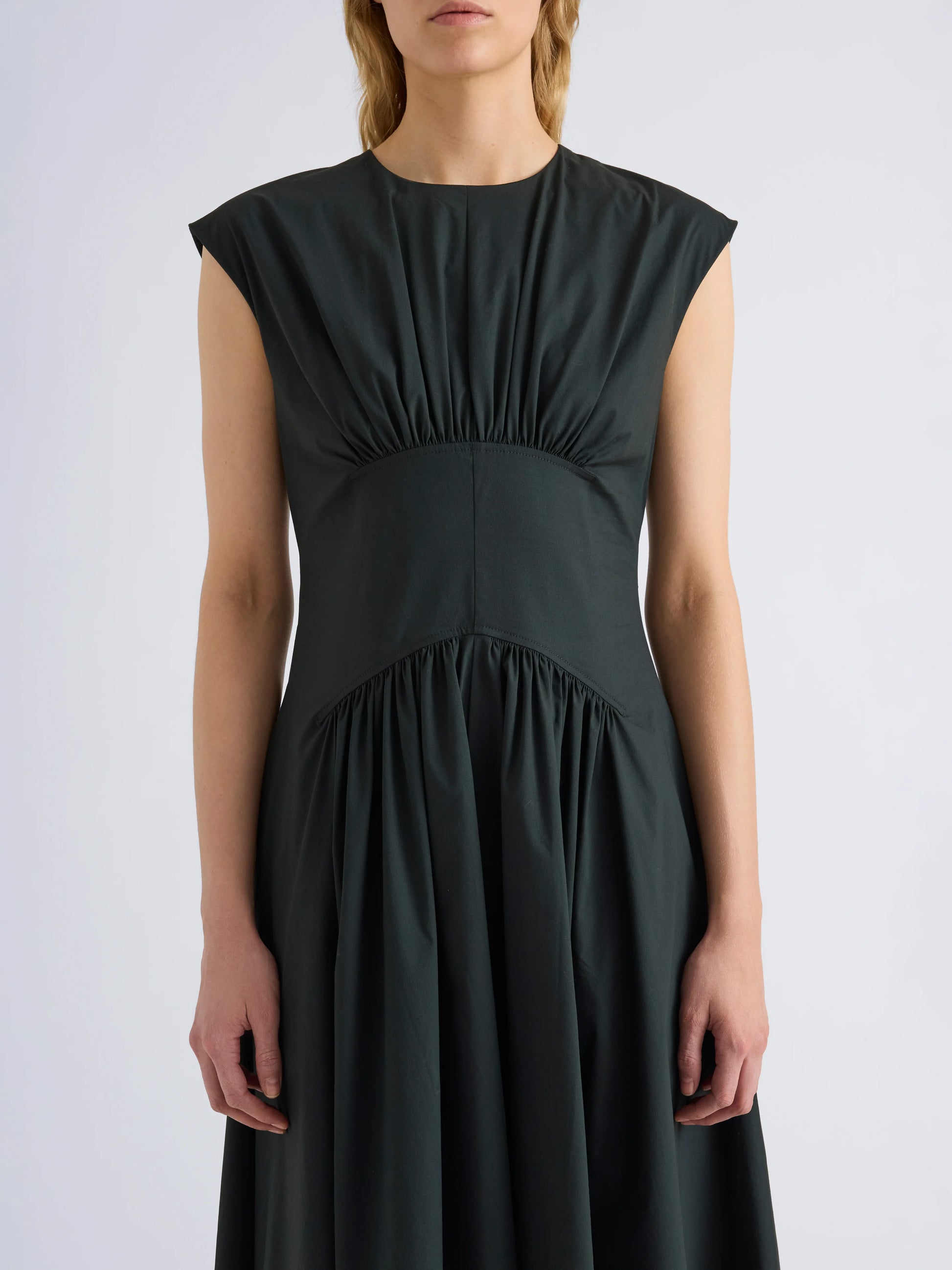 Wearing the Proenza Schouler White Label Nyssa Stretch Cotton Dress, a sleeveless dark green women's dress featuring gathered fabric details at the shoulders, waist, and skirt, the person stands against a plain light background.
