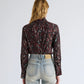 A person with wavy, light brown hair is seen from the back, wearing a maroon Ansel Printed Soft Twill Top by Proenza Schouler White Label with a subtle pattern, tucked into high-waisted light blue jeans, set against a plain background.