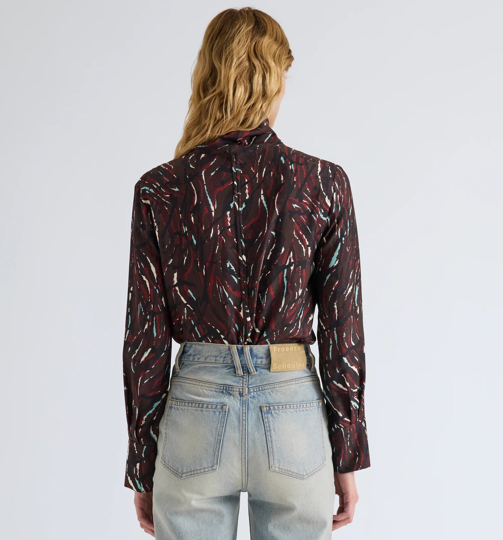 A person with wavy, light brown hair is seen from the back, wearing a maroon Ansel Printed Soft Twill Top by Proenza Schouler White Label with a subtle pattern, tucked into high-waisted light blue jeans, set against a plain background.