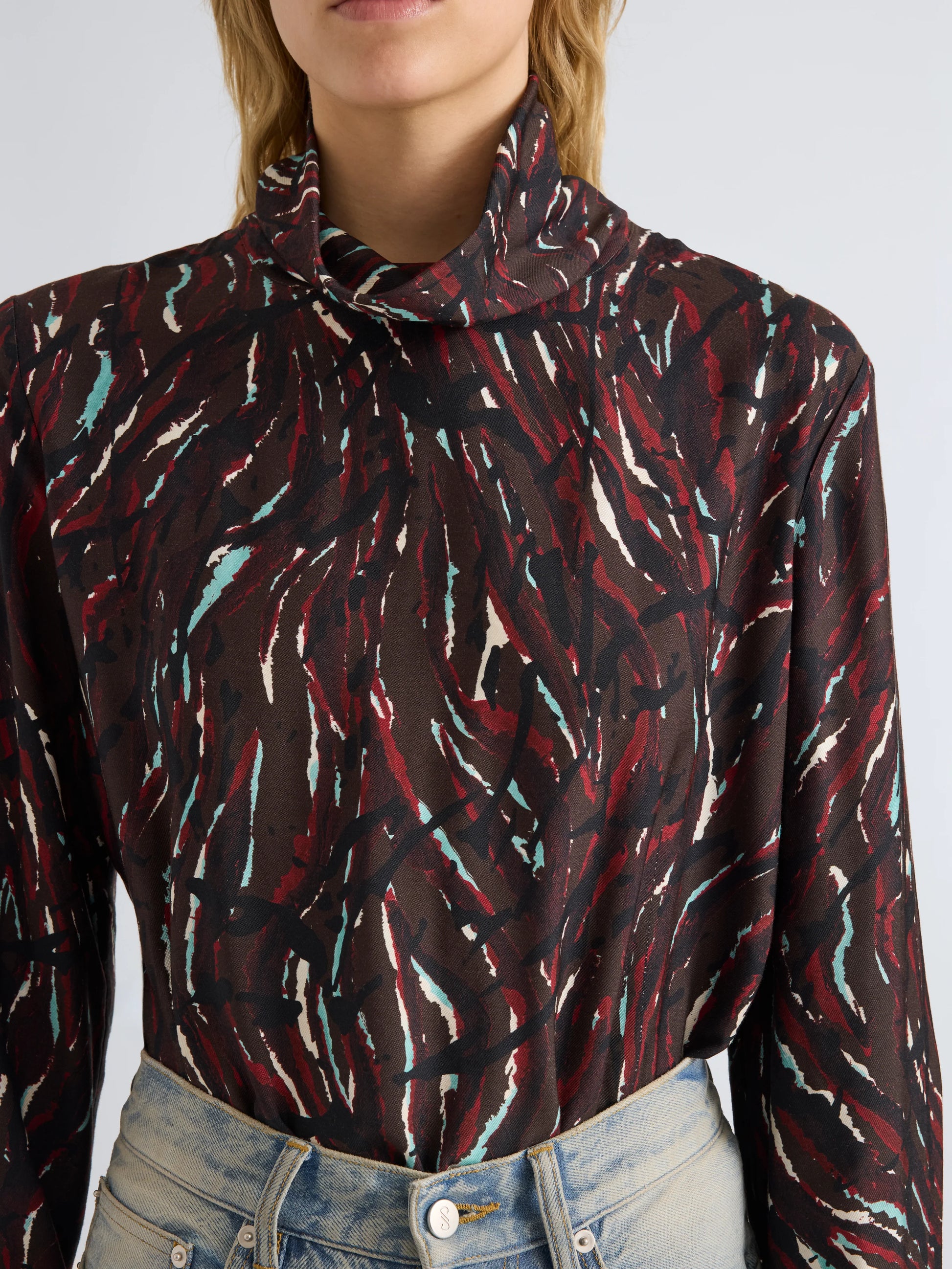 A person wearing the Proenza Schouler White Label Ansel Printed Soft Twill Top—a long-sleeve, high-neck piece featuring an abstract maroon, white, and teal pattern with a subtle cowl turtleneck—paired with light blue jeans.