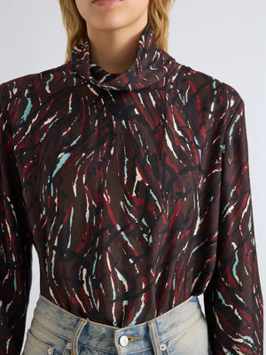 A person wearing the Proenza Schouler White Label Ansel Printed Soft Twill Top—a long-sleeve, high-neck piece featuring an abstract maroon, white, and teal pattern with a subtle cowl turtleneck—paired with light blue jeans.